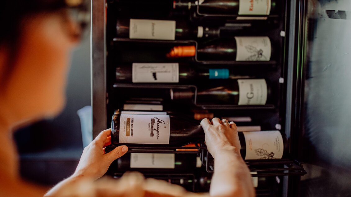 Eine Person greift nach einer Flasche Wein aus einem Weinregal. Das Regal enthält verschiedene Weinflaschen mit unterschiedlichen Etiketten. | © Karin Bergmann
