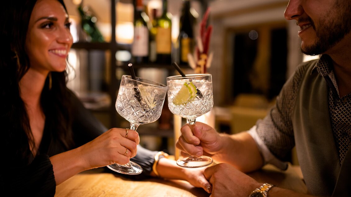 Ein Paar stoßt mit zwei Gin tonic Cocktails auf einem Tisch an. Im Hintergrund sind Weinflaschen und eine gemütliche Atmosphäre zu sehen. | © Karin Bergmann