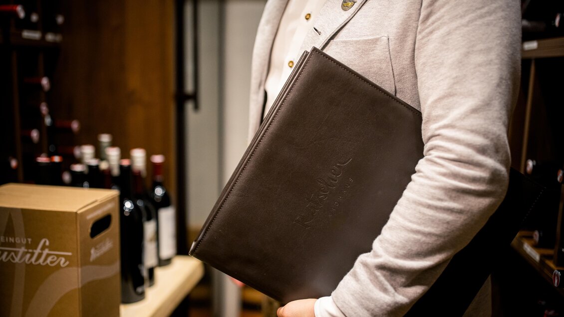 Eine Person hält eine dunkle Ledermappe, die Weinkarte, in der Hand. Im Hintergrund sind Weinflaschen und eine Kiste zu sehen. | © Karin Bergmann