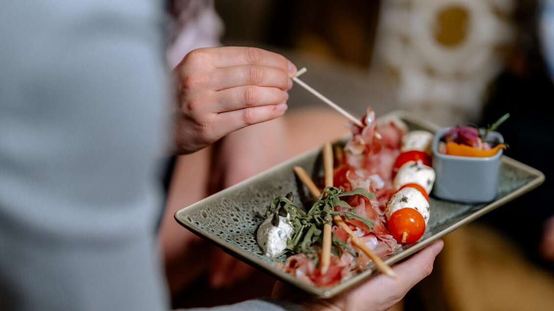 Ein Teller mit Snacks, darunter Kirschtomaten, Mozzarella und Schinken. Im Hintergrund sind weitere Speisen und eine Person zu sehen, die mit einem Zahnstocher anrichtet. | © Obersberger