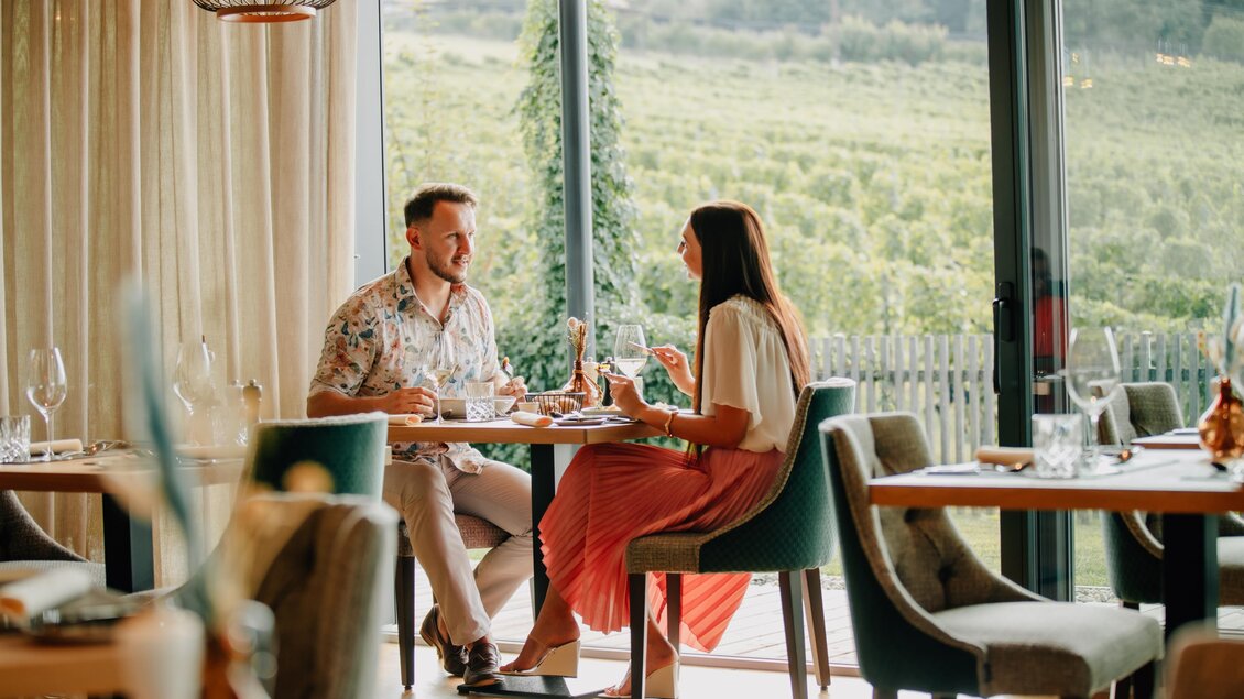 Ein Paar sitzt an einem Tisch in einem modernen Restaurant mit Blick auf die Weinberge. Sie genießen ihr Essen und sprechen miteinander. | © Karin Bergmann