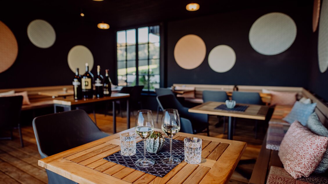 Ein moderne Loungebereich mit offener Terrasse und Blick in die Weingärten, mit stilvollen Möbeln und einer gemütlichen Atmosphäre. Auf einem Tisch stehen Weingläser und Dekoration, während im Hintergrund Flaschen platziert sind. Das ganze ist überdacht | © Karin Bergmann