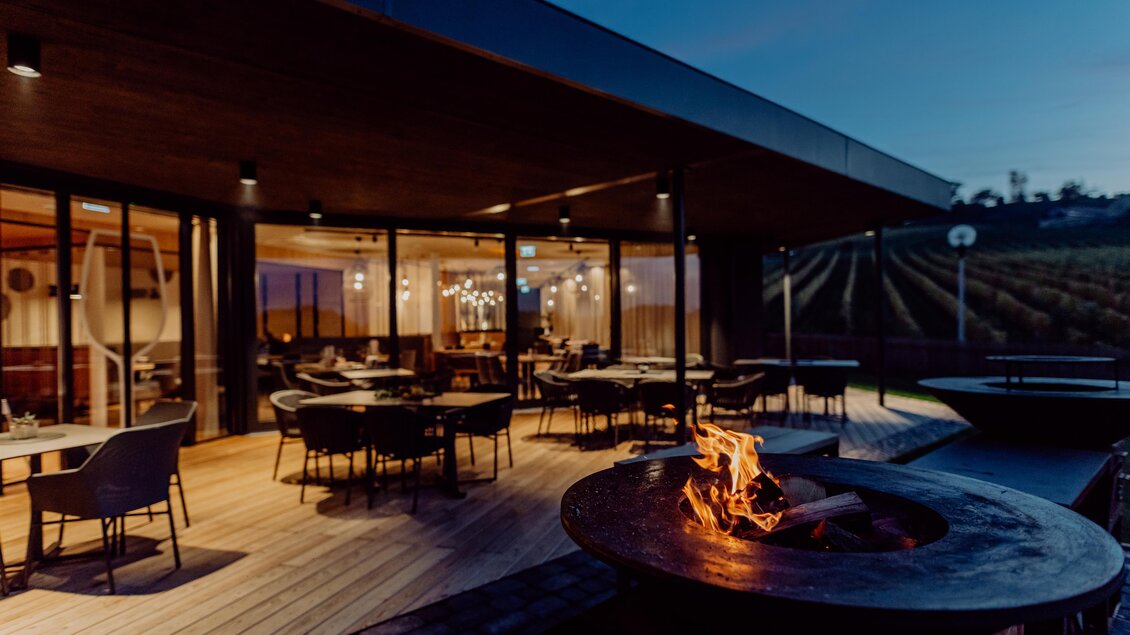 Abendamtosphäre im modernen Restaurant mit einer schönen Terrasse und Blick auf Weinreben. Der gemütliche Außenbereich ist mit einem Feuerschale ausgestattet und wird von sanftem Licht beleuchtet. | © Karin Bergmann