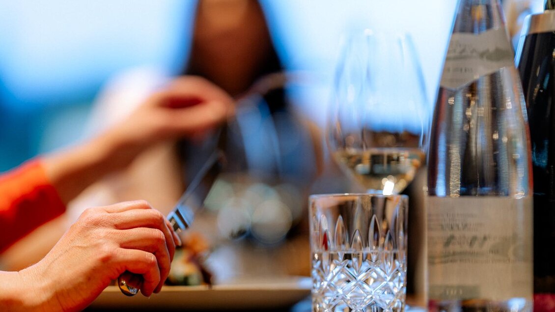 Ein gedeckter Tisch mit einem Glas Wasser und einem Glas Weißwein. Im Vordergrund wird eine Mahlzeit mit Besteck serviert. alles sehr elegant und modern | © Obersberger