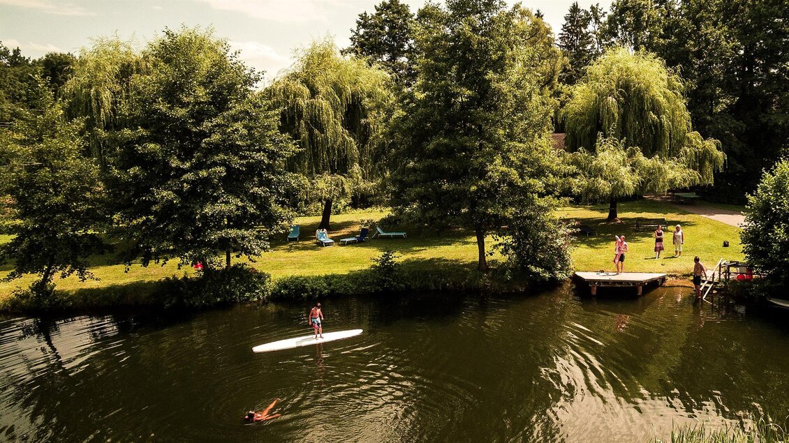 Ein ruhiger Gewässerbereich mit Schwimmern und Stand-Up-Paddle-Boardern. Umgeben von grünen Bäumen und einer einladenden Wiese. | © Rauch-Hof