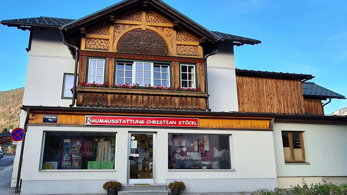 Ein traditionelles Gebäude mit einem kunstvoll geschnitzten Dachüberstand. Im Erdgeschoss befindet sich ein Geschäft mit der Aufschrift "Raumausstattung Christian Stöckl". | © Stephanie Bor