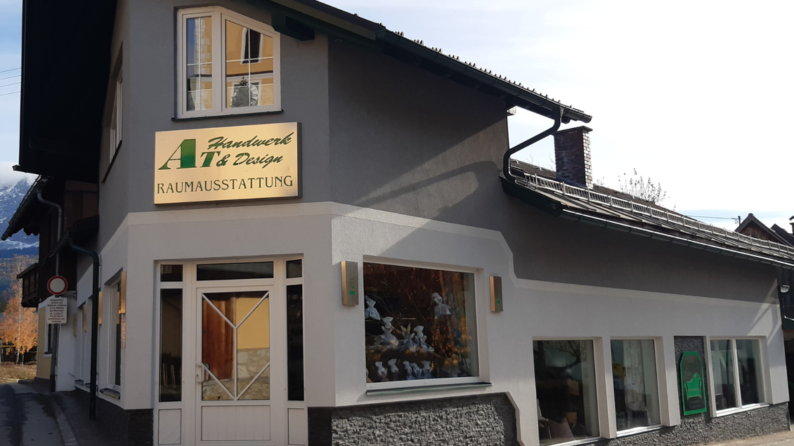 Ein Geschäft mit dem Namen „AT Handwerk & Design“ in einem zweistöckigen Gebäude. Die Fassade ist hellgrau und gut gepflegt, mit großen Fenstern, die einen einladenden Eindruck hinterlassen. | © TVB Ausseerland Salzkammergut_Daniela Casari