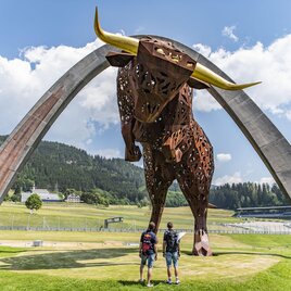 A large, impressive metal sculpture of a bull stands under an arch. Two people are fascinatedly looking at the art installation in a green landscape. | © Erlebnisregion Murtal