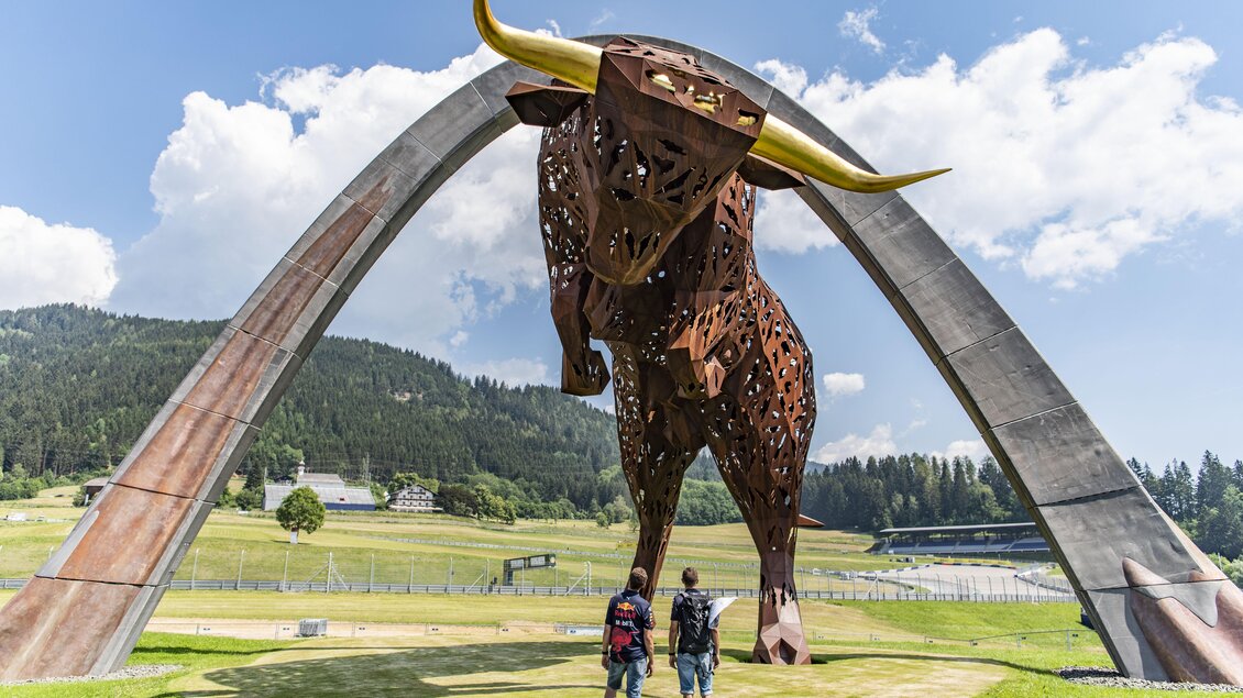 Eine große, beeindruckende Metallskulptur eines Bullen steht unter einem Bogen. Zwei Personen betrachten fasziniert die Kunstinstallation in einer grünen Landschaft. | © Erlebnisregion Murtal