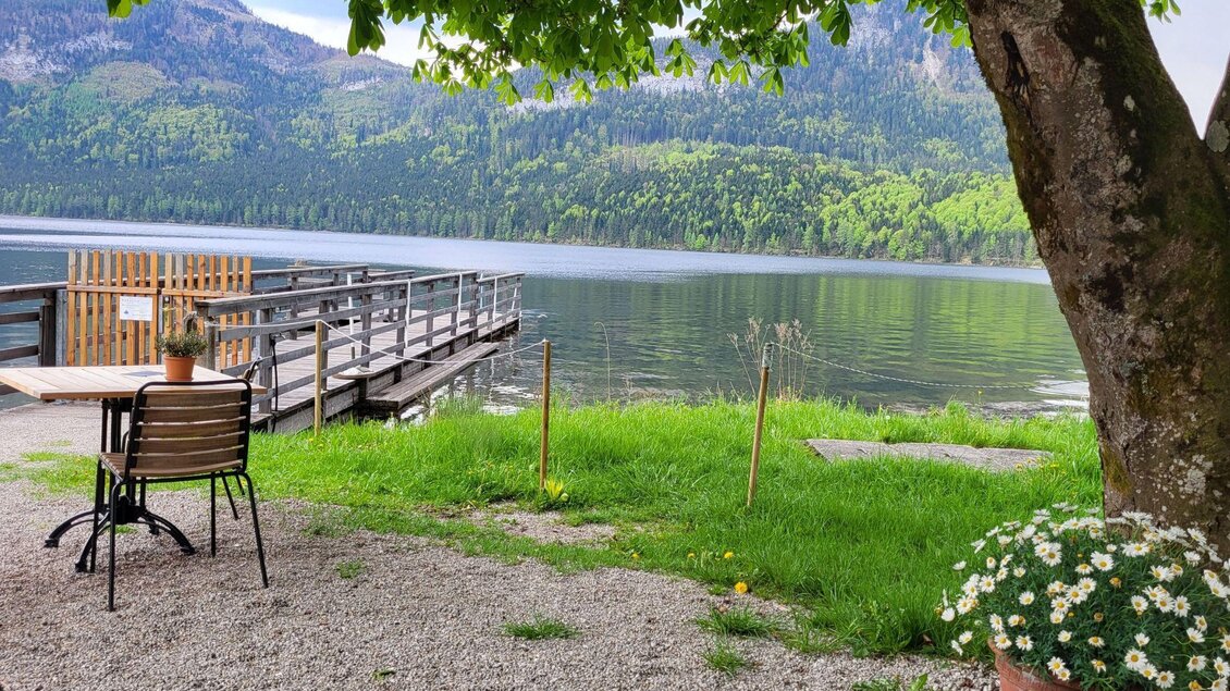Ein ruhiger See mit grünen Bergen im Hintergrund. Am Ufer steht ein Tisch mit Stühlen und Blumen in einem Topf. | © Petra Kirchschlager