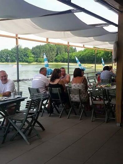 Ein Restaurant auf einer Terrasse am Wasser mit Gästen, die entspannt essen und trinken. Sonnenschirme schützen vor der Sonne und die Umgebung ist grün und einladend. | © Reiners am Planksee