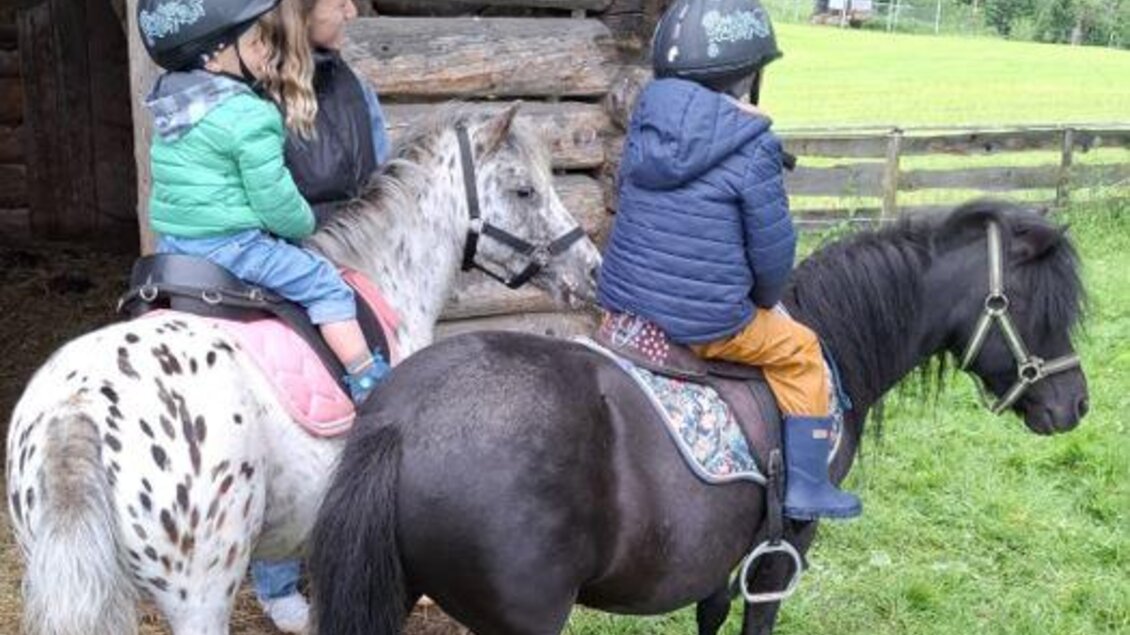 Zwei Kinder reiten auf Ponys in einer grünen Wiese. Im Hintergrund ist eine Holzstruktur und eine schöne Landschaft zu sehen. | © Hirzhof
