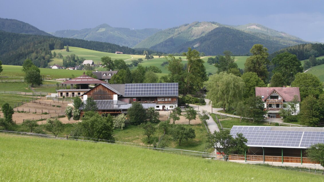 Eine malerische Landschaft mit einem Bauernhof und hügeligen Wiesen. Im Hintergrund sind Berge und ein bewölkter Himmel zu sehen. | © Anton Stockner