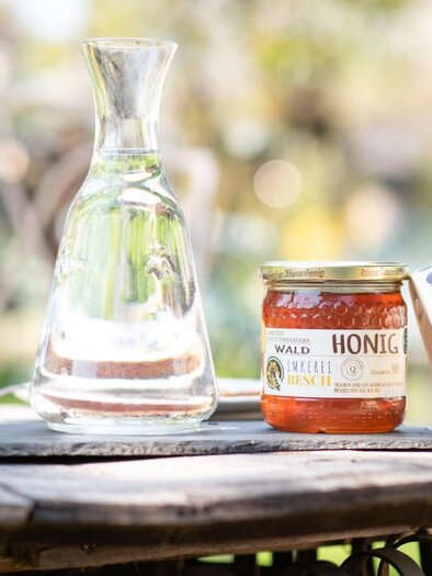 A table with a decorative cup, a glass carafe, and a honey jar. A dotted ribbon lies alongside, adding a charming touch to the arrangement. | © RM SW GmbH | webquartier.at