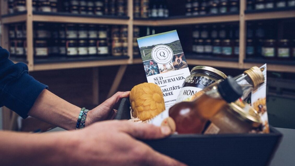 Ein Korb mit regionalen Produkten, darunter ein Brot, Getränke und Informationsmaterial. Im Hintergrund befinden sich Regale mit eingemachten Lebensmitteln. | © RM SW GmbH | webquartier.at