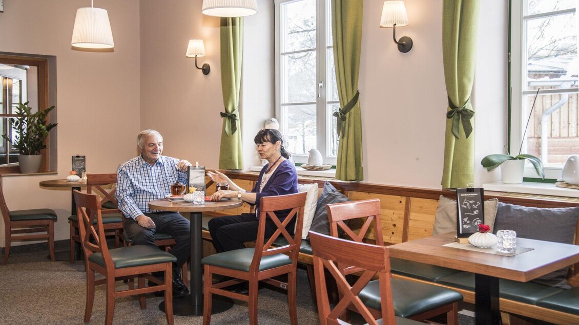 Ein gemütliches Café mit Holzstühlen und grünen Vorhängen. Ein älteres Paar sitzt zusammen am Tisch und genießt Getränke. | © Jufa Hotels
