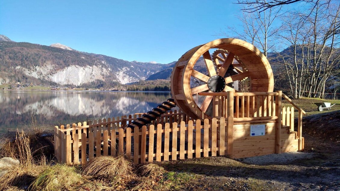 Ein Holz-Wasserrad steht direkt am Ufer eines ruhigen Sees. Im Hintergrund sind Berge und ein klarer blauer Himmel zu sehen. | © Jufa Hotels
