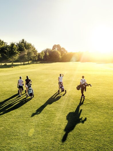 Vier Golfer gehen über einen weiten Rasenplatz bei Sonnenuntergang. Die lange Schatten und das goldene Licht schaffen eine entspannte Atmosphäre. | © AdobeStock_123241965