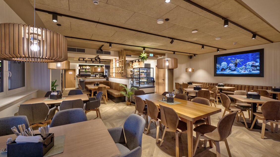 Ein modernes Restaurant mit Holzmöbeln und stilvollen Lampen. An der Wand befindet sich ein großer Bildschirm mit einem Aquarienbild. | © Gross Genuss GmbH