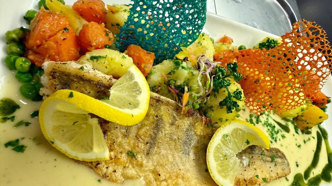 Ein kunstvoll angerichteter Teller mit frischem Fisch, umgeben von bunten Gemüsewürfeln und dekorativen Chips. Zitronenscheiben verleihen dem Gericht einen frischen Akzent. | © Gross Genuss GmbH