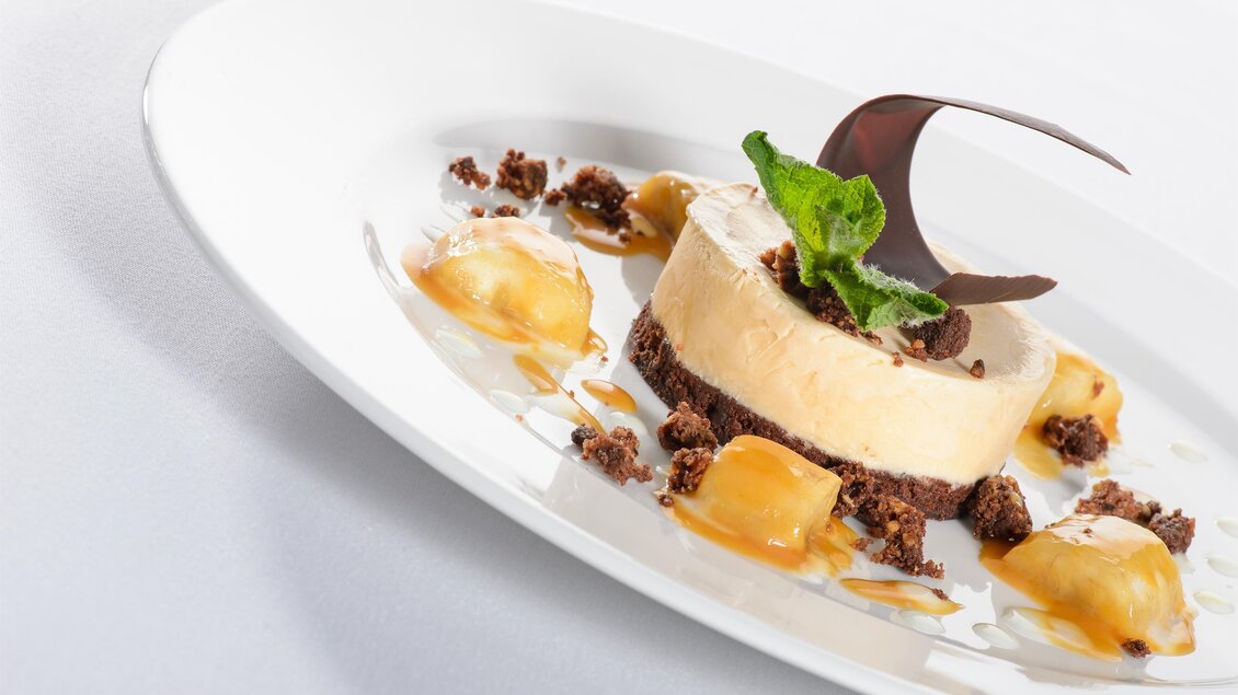 Ein eleganter Dessertteller mit einer Käsecreme, dekoriert mit Schokolade und frischer Minze. Umgeben von kleinen Fruchtstücken und Keksbröseln. | © Tauroa GmbH