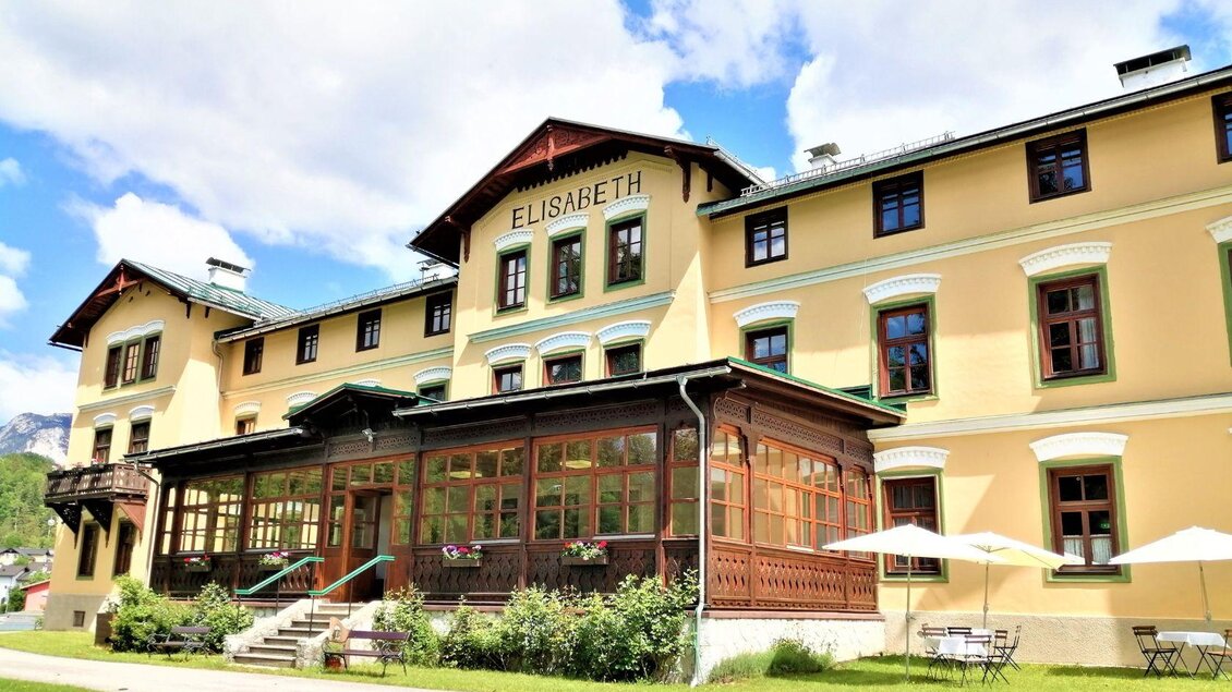 Ein historisches Hotel mit gelber Fassade und einer schönen Veranda. Umgeben von grüner Landschaft und mit Tischen im Freien. | © Gasthof Elisabeth