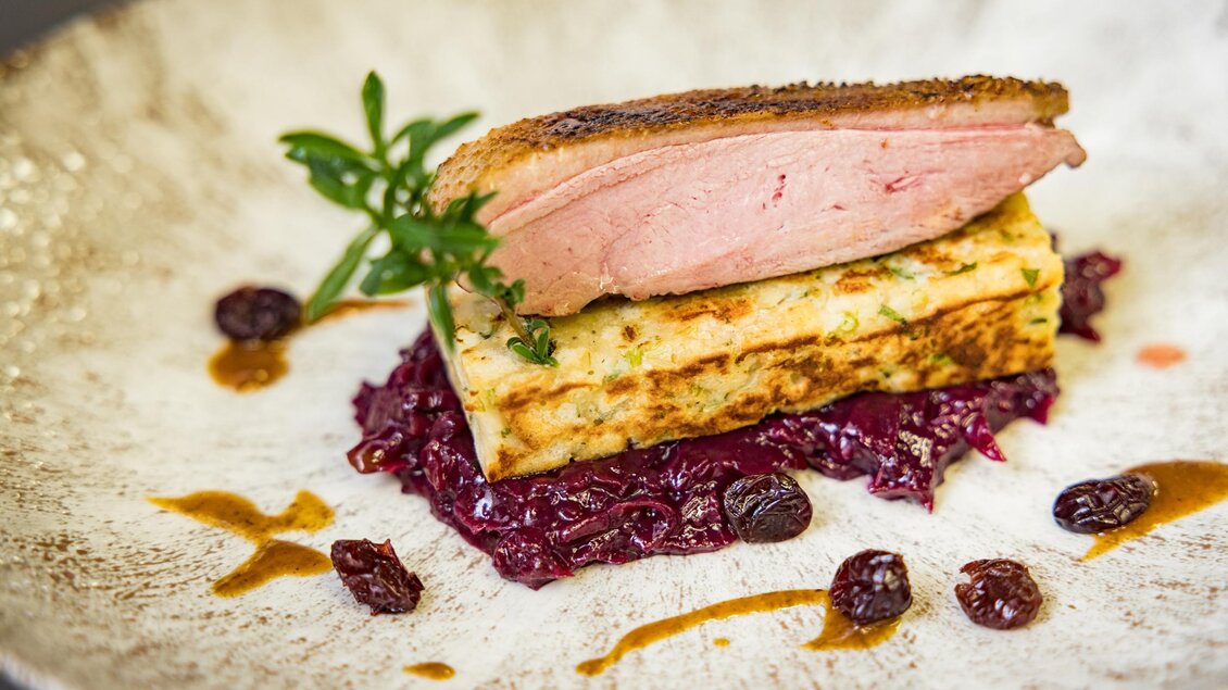 Ein ansprechend angerichteter Teller mit gebratenem Fleisch auf einer Schicht aus Kartoffelpüree und Rotkraut. Die Dekoration umfasst einige Kräuter und kleine Preiselbeeren. | © Netzwerk Kulinarik wildbild