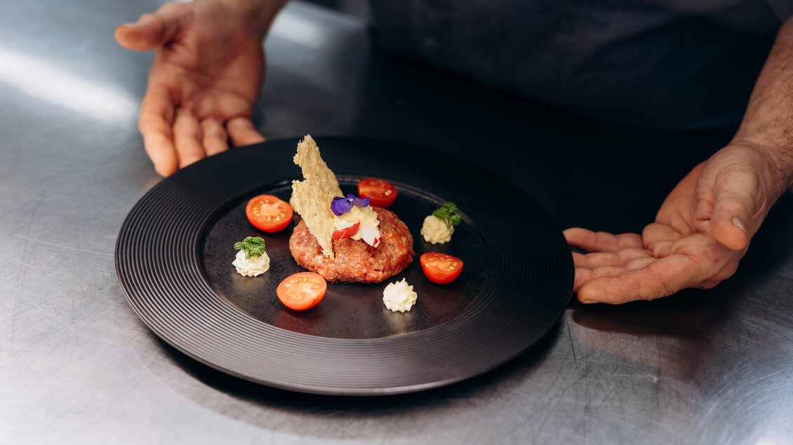 Ein kunstvoll angerichteter Teller mit einer Fleischscheibe, Kirschtomaten und verschiedenen Beilagen. Der Koch präsentiert das Gericht mit beiden Händen vor sich.