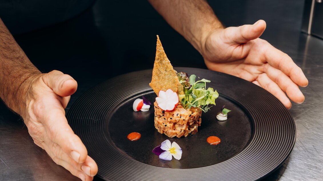 Ein kunstvoll angerichteter Teller mit einem kleinen Gericht. Der Koch präsentiert das Essen mit zwei Händen, um die Details hervorzuheben.