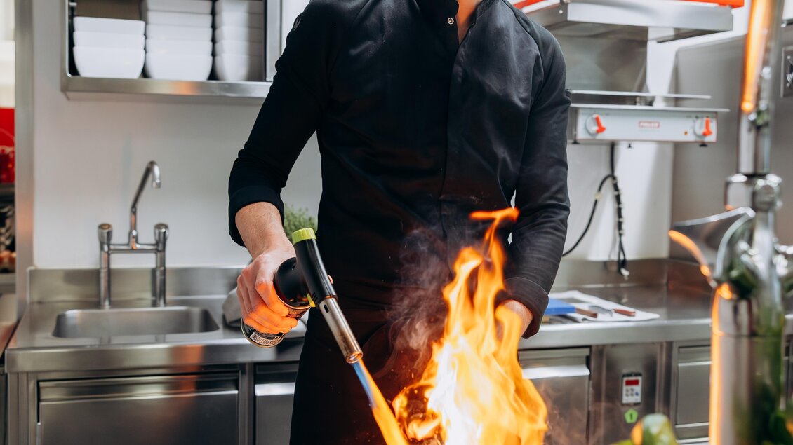 Ein Koch zündet in einer modernen Küche mit einer Flamme eine Pfanne an. Die Umgebung ist sauber und gut organisiert, ideal für die Zubereitung von Speisen.