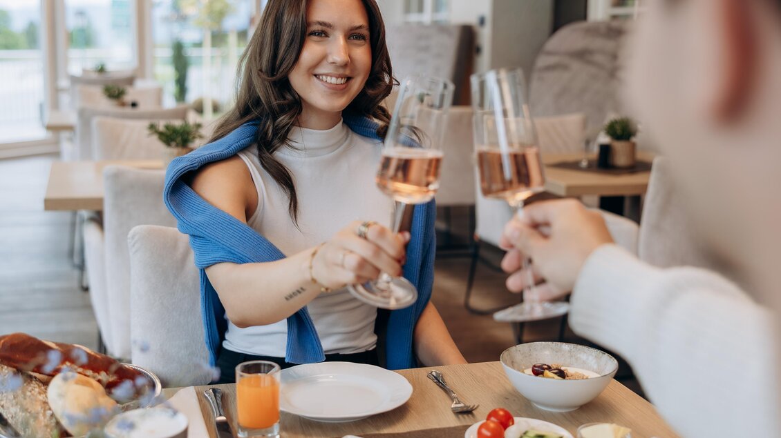 Ein fröhliches Frühstück in einem modernen Restaurant. Eine Frau stößt mit einem Glas Sekt an, während der Tisch mit verschiedenen Köstlichkeiten gedeckt ist.