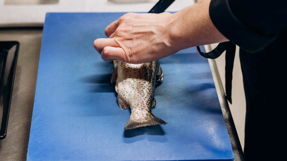 Eine Person schneidet einen Fisch auf einem blauen Schneidebrett in einer modernen Küche. Im Hintergrund liegen weitere Fische auf einem Blech.