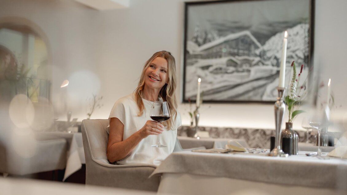 Eine Frau sitzt in einem eleganten Restaurant und lächelt, während sie ein Glas Wein hält. Der Tisch ist geschmackvoll mit Kerzen und Blumen dekoriert. | © Vison