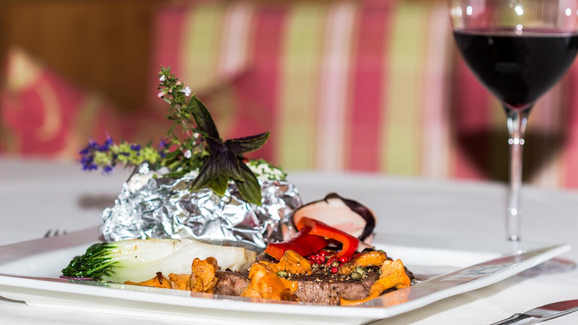 Ein elegant angerichtetes Gericht mit Gemüse, Fleisch und Kräutern auf einem weißen Teller. Im Hintergrund steht ein Glas Rotwein und die Tischdekoration hat sanfte Farben. | © caro lang