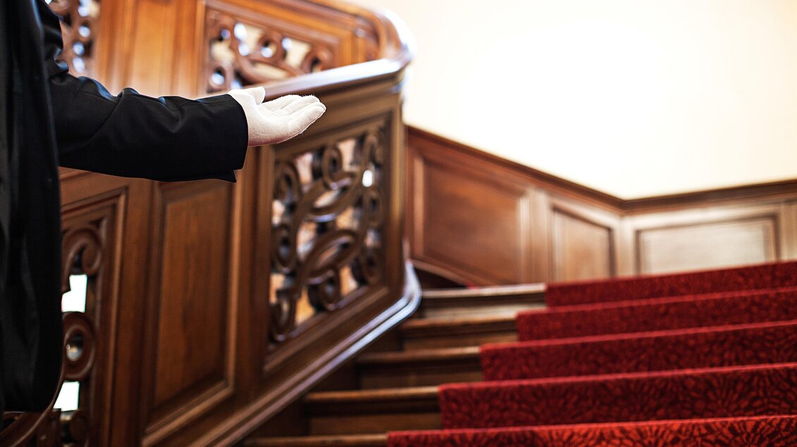 Ein eleganter Treppenaufgang mit rotem Teppich und kunstvoller Holzhandlauf. Eine Person weist freundlich auf die Treppe hin.