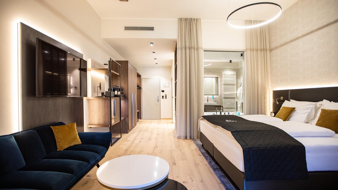 Ein modernes Hotelzimmer mit einem komfortablen Bett und einem eleganten Sofa. Die helle Einrichtung und die stilvolle Beleuchtung schaffen eine einladende Atmosphäre.