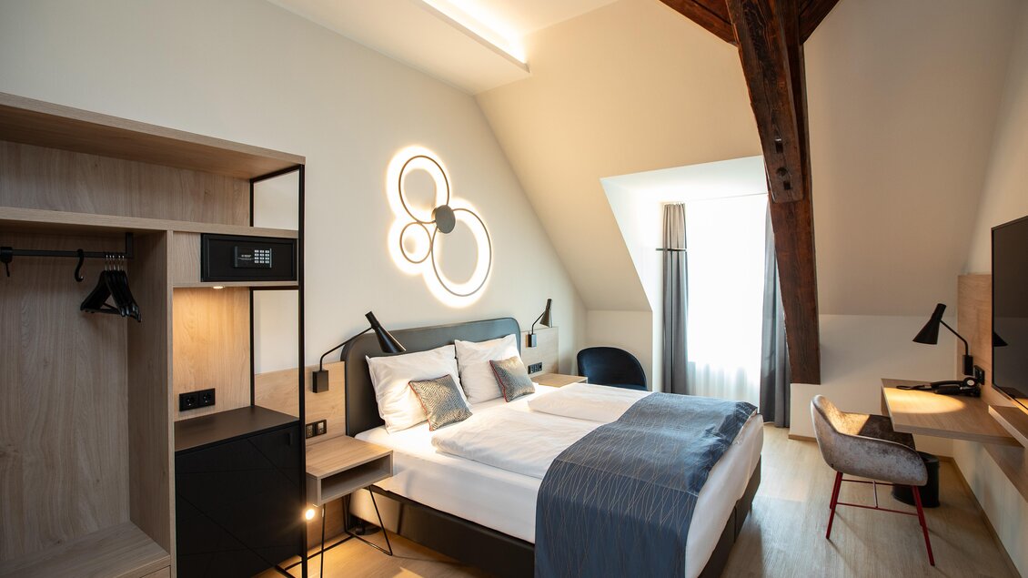 Ein modernes Hotelzimmer mit einem großen Bett und stilvollen Möbeln. Die große Fensterfront sorgt für viel natürliches Licht und eine angenehme Atmosphäre.