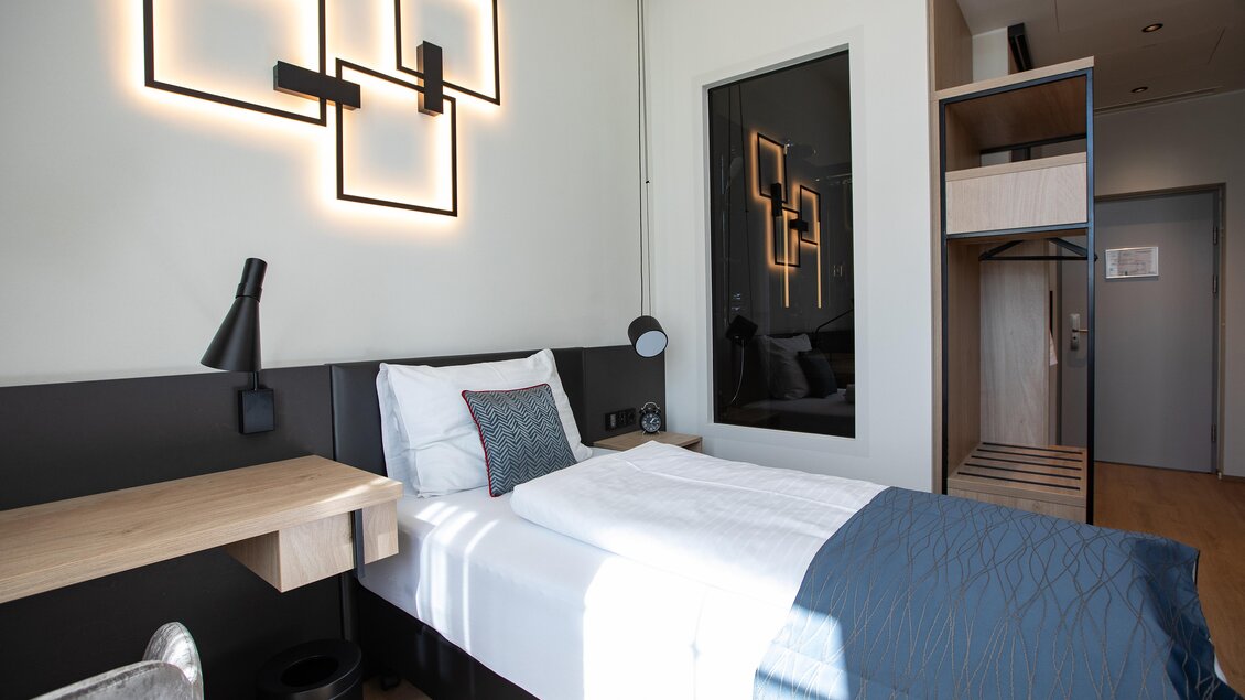 Ein modernes Hotelzimmer mit einem Einzelbett und elegantem Dekor. Die Wände sind hell, und es gibt eine gemütliche Arbeitsfläche sowie einen Kleiderschrank.