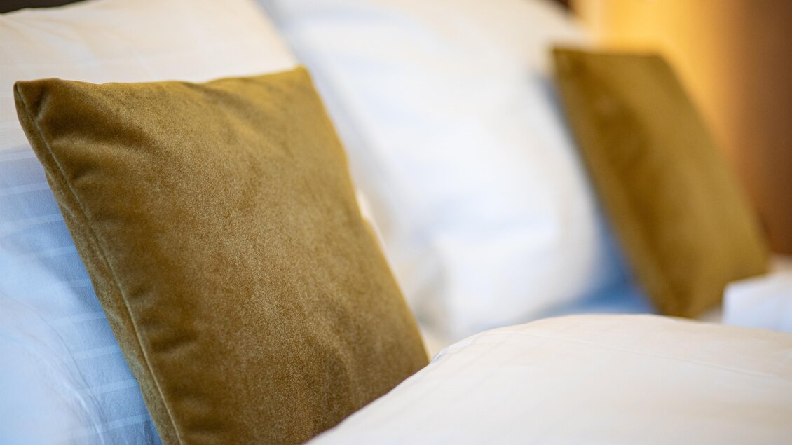 Ein gemütliches Bett mit weißen Kissen und zwei braunen Dekokissen. Die warme Atmosphäre lädt zum Entspannen ein.