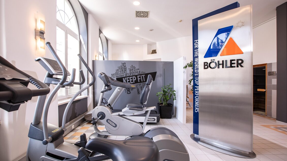 Ein moderner Fitnessraum mit verschiedenen Trainingsgeräten wie Crosstrainer und Fitnessbikes. Im Hintergrund steht ein Schild mit dem Logo von Böhler.