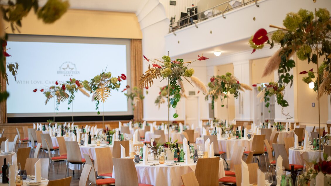 Ein festlich dekorierter Saal mit eleganten Tischen und Blumenarrangements. Im Hintergrund ist eine Präsentationsfläche zu sehen.