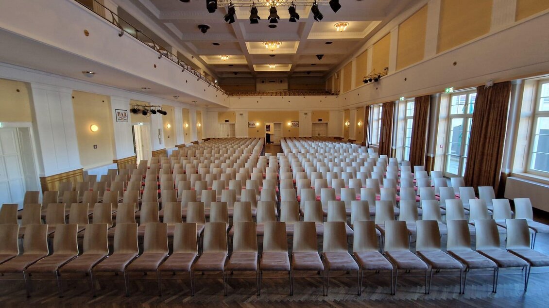 Ein großer Saal mit Reihen von leeren Stühlen. Die Wände sind hell und die Beleuchtung ist warm.