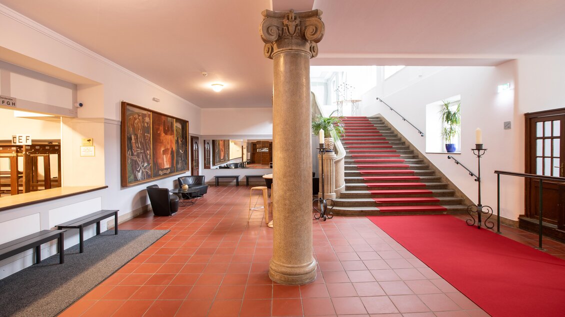 Ein heller Empfangsbereich mit einem roten Teppich und einer Treppe im Hintergrund. An der Wand hängen große Gemälde und es gibt gemütliche Sitzgelegenheiten.