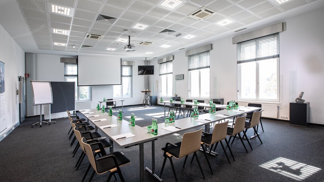 Ein moderner Besprechungsraum mit einem langen Tisch und Stühlen. Es gibt Fenster, die viel Tageslicht hereinlassen, und die Ausstattung ist schlicht und funktional.