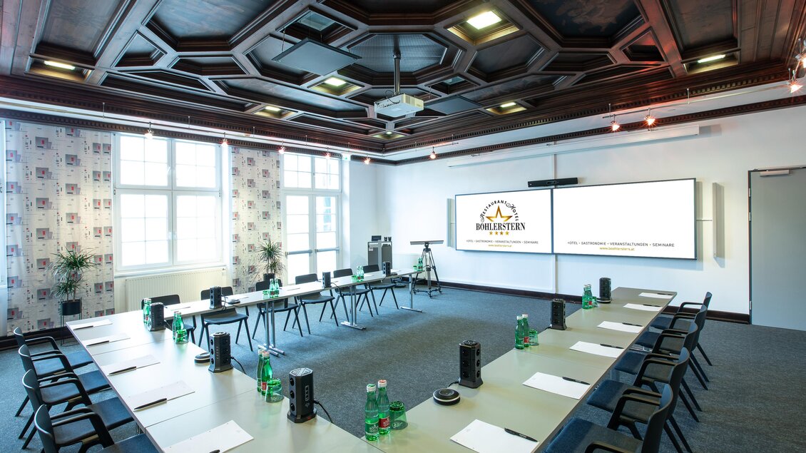 Ein modernes Konferenzzimmer mit einem länglichen Tisch und schwarzen Stühlen. An der Wand sind zwei große Bildschirme sowie Fenster mit viel natürlichem Licht.