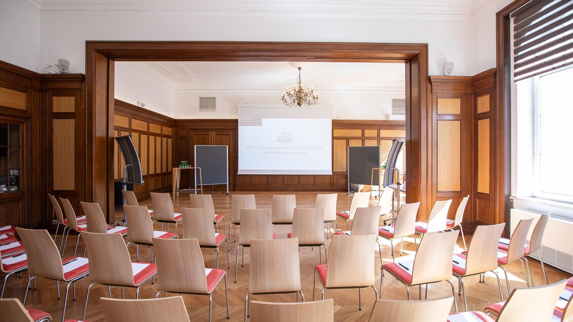 Ein Konferenzraum mit Holzverkleidung und mehreren Stühlen in Reihen. In der Mitte steht ein Projektor mit einem vorbereiteten Bildschirm.