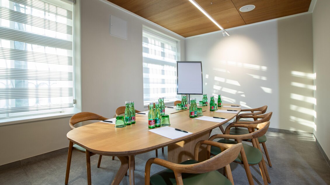 Ein moderner Besprechungsraum mit einem großen Holztisch und Stühlen. Auf dem Tisch stehen Mineralwasserflaschen und Notizblätter, während das Licht durch die Fenster strömt.