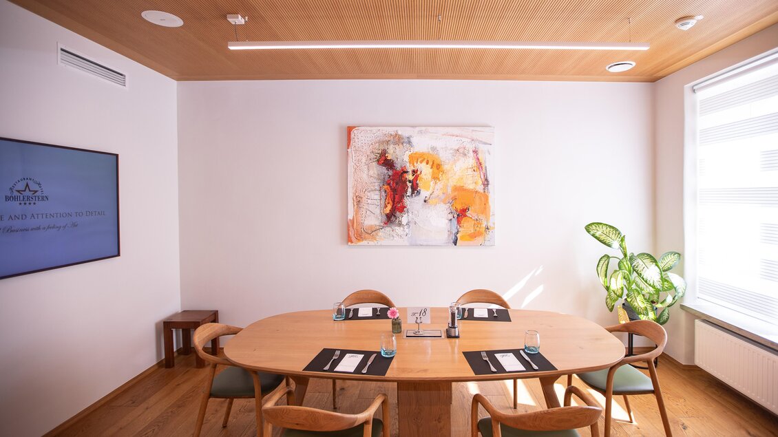 Ein modernes Konferenzzimmer mit einem großen Holztisch und sechs Stühlen. An der Wand hängt ein farbenfrohes Kunstwerk, und ein großer Bildschirm ist ebenfalls sichtbar.