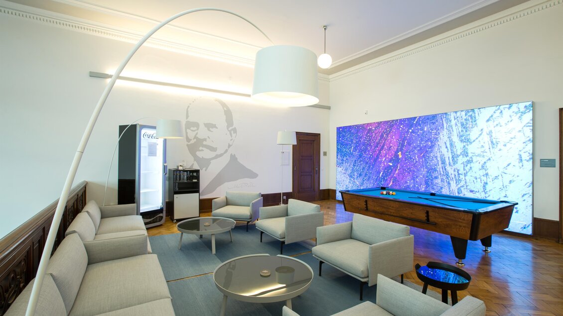 Ein modernes Wohnzimmer mit einer eleganten Couchgarnitur und einem Billardtisch. An der Wand befindet sich ein großer Bildschirm mit bunten grafischen Mustern.