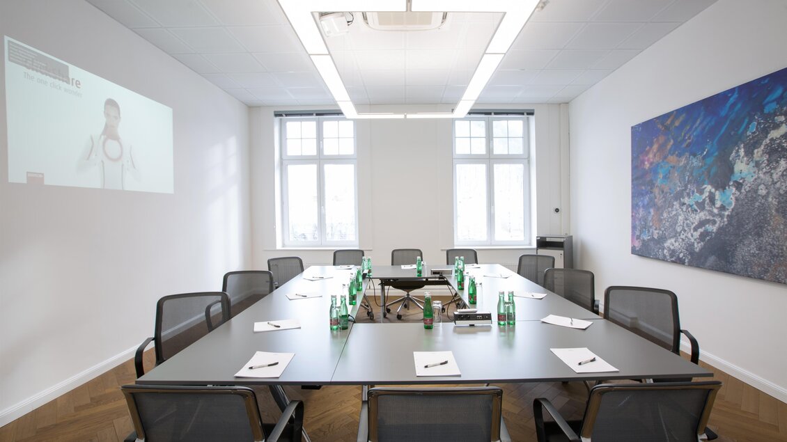 Ein moderner Besprechungsraum mit einem großen Tisch und mehreren Stühlen. Auf dem Tisch befinden sich Wasserflaschen und Notizblätter, während eine Präsentation an der Wand angezeigt wird.